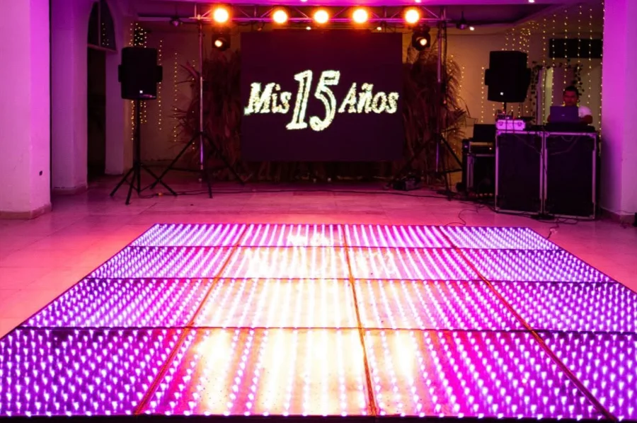 Pista de baile LED iluminada frente a una pantalla grande con el mensaje Mis 15 Años, parlantes y cabina de DJ